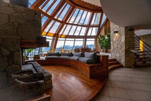 A spacious living area with a stone fireplace, a curved seating area, and a view of mountains through large windows. - Sol Arrayan Hotel & Spa (Villa La Angostura)
