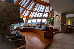 Amplia sala de estar con chimenea de piedra, zona de estar curva y vistas a montañas a través de grandes ventanales.