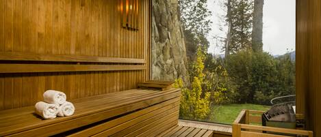 Una sauna de madera con bancos y un asiento de banco, una toalla sobre el banco, y vistas a árboles y una montaña.