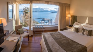 A hotel room with a large bed, a desk, a chair, a nightstand, and a view of a lake and mountains. - Sol Arrayan Hotel & Spa (Villa La Angostura)