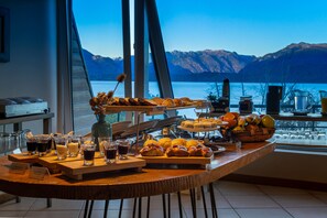 A breakfast buffet with pastries, fruits, and beverages on a wooden table with a view of mountains and a lake. - Sol Arrayan Hotel & Spa (Villa La Angostura)