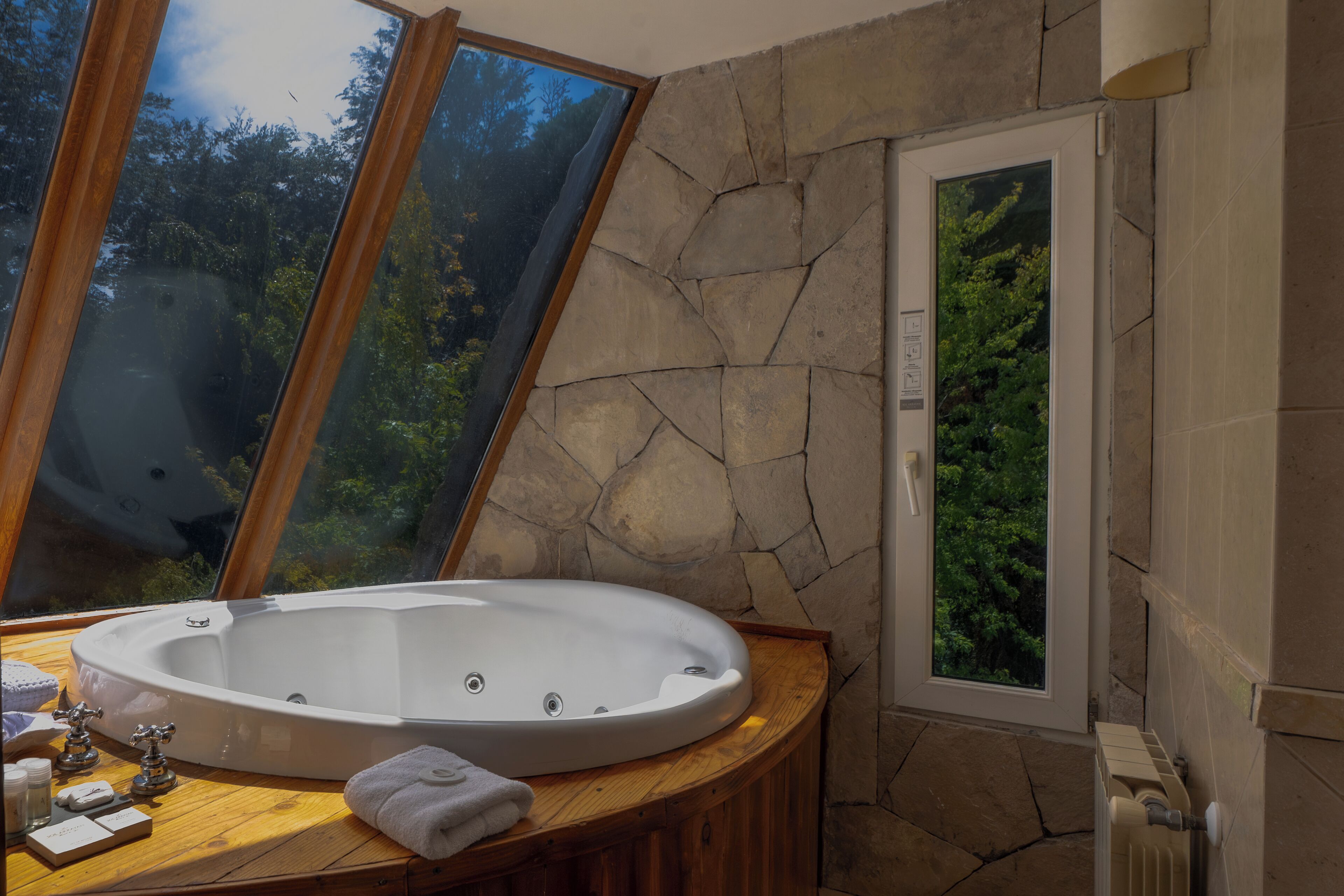 Ein Badezimmer mit Steinwand, Holzwaschtisch, einer großen Badewanne und einem Fenster mit Blick auf Bäume.
