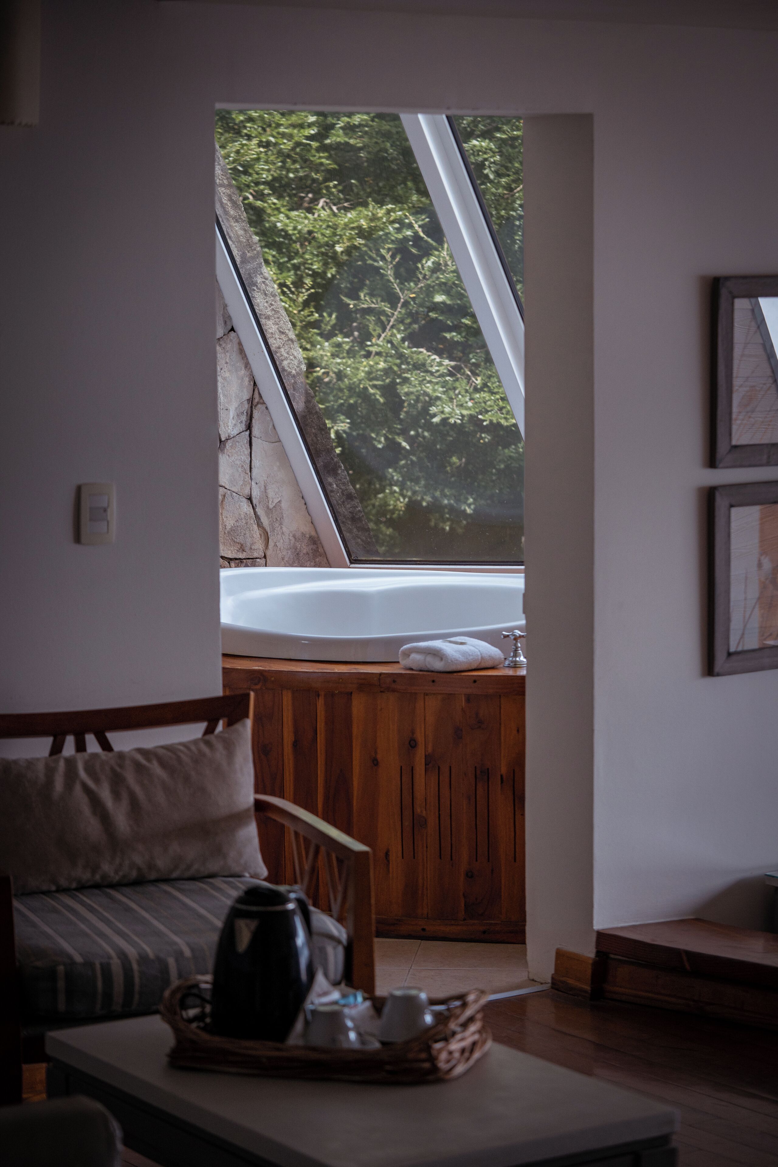 A room with a wooden bench, a table with a kettle, a bathtub, and a view of greenery through a window.