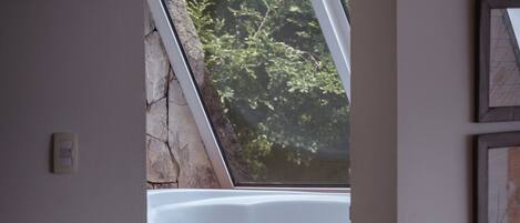 A room with a wooden bench, a table with a kettle, a bathtub, and a view of greenery through a window.