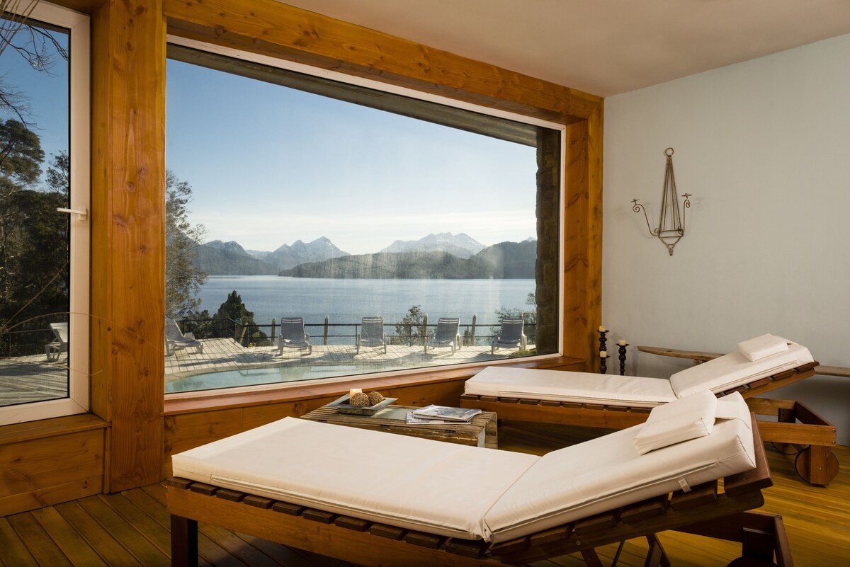 Una habitación con dos camas de madera, un amplio ventanal con vistas a un lago y montañas, y una mesita con una vela y un libro.