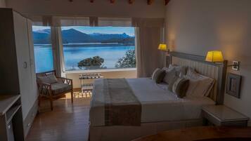 A bedroom with a bed, bedside lamps, a chair, and a view of a lake and mountains.