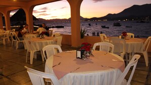 Un área de comedor al aire libre con mesas y sillas, con vistas a una masa de agua con barcos y un atardecer de fondo.
