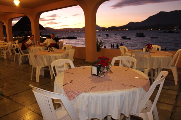 An outdoor dining area with tables and chairs, overlooking a body of water with boats and a sunset in the background.