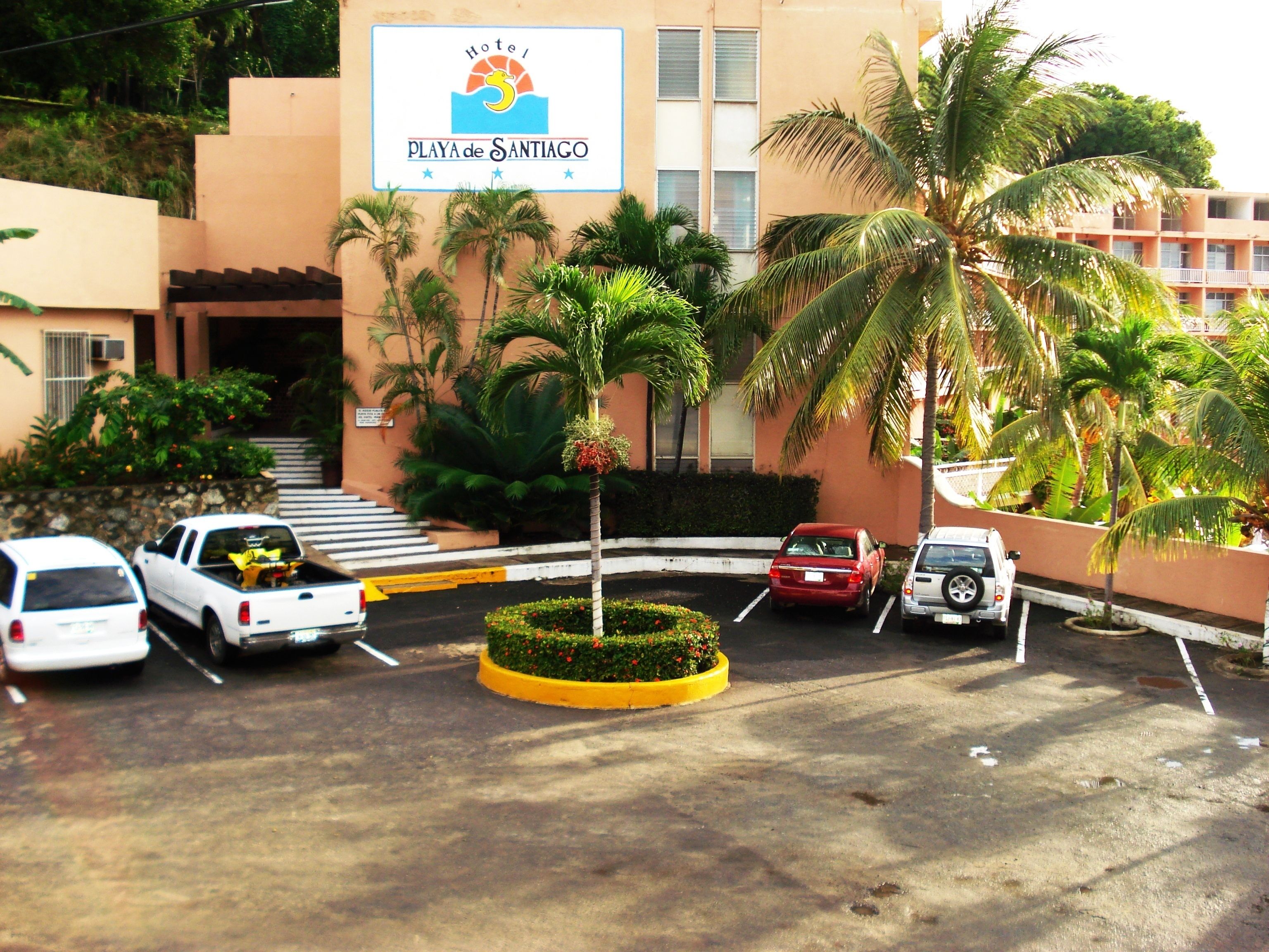Un hotel con un cartel que dice "Hotel Playa de Santiago", que cuenta con un estacionamiento con varios coches, palmeras y un jardín bien cuidado.