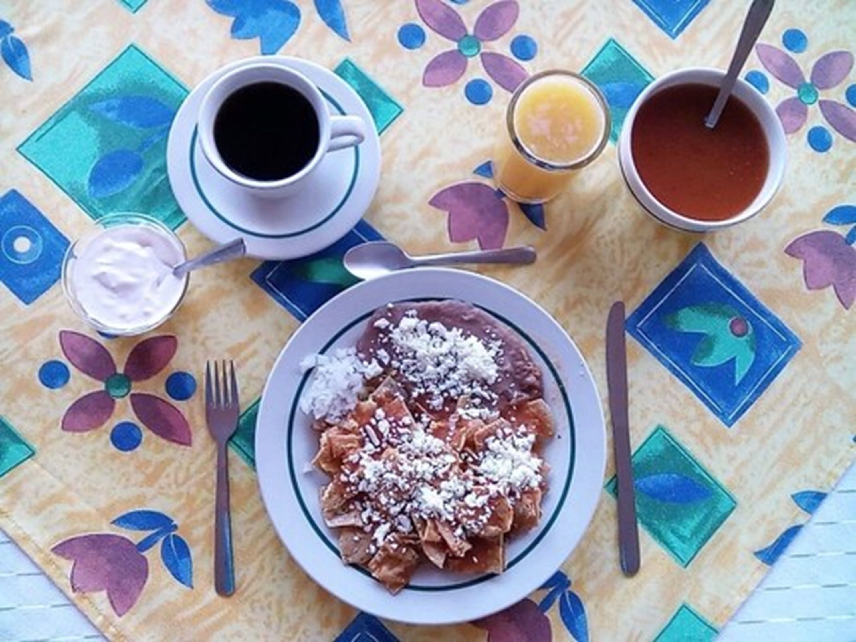 A table with a colorful tablecloth, a plate of enchiladas, a cup of coffee, a glass of orange juice, and a glass of red sauce.