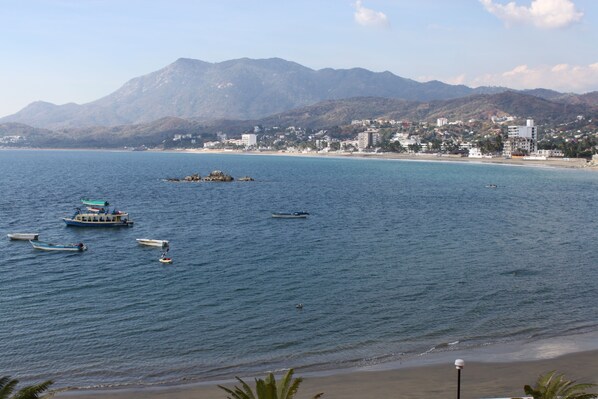 A coastal view with boats on the water, a beach, and a mountain range in the background.