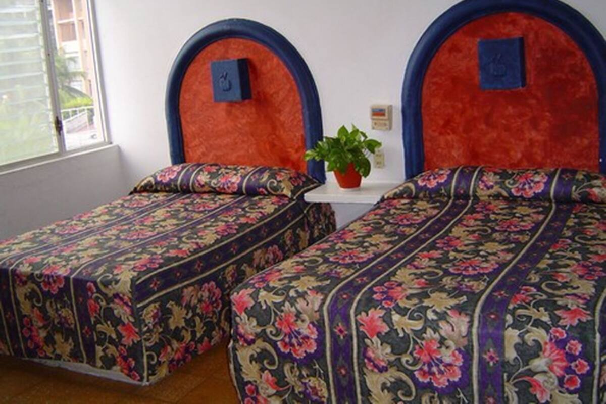 Ein Zimmer mit zwei Betten, jeweils mit einem blumigen Überwurf und passenden Kissen. Dazwischen steht ein kleiner Tisch mit einer Topfpflanze.