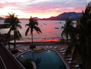 Vista a la piscina con sillones de descanso y palmeras, con vista a un cuerpo de agua y montañas al atardecer.