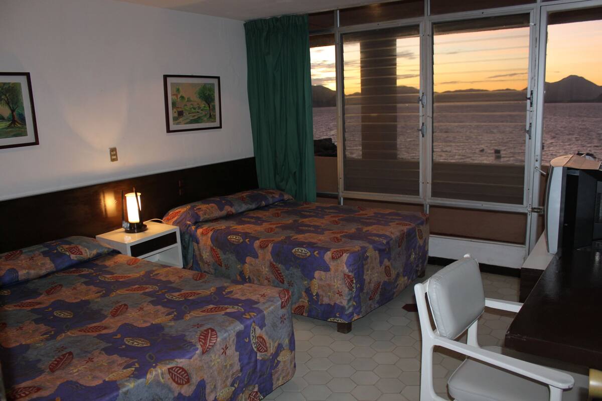 Ein Hotelzimmer mit zwei Betten, einem Schreibtisch und Blick auf das Meer bei Sonnenuntergang.