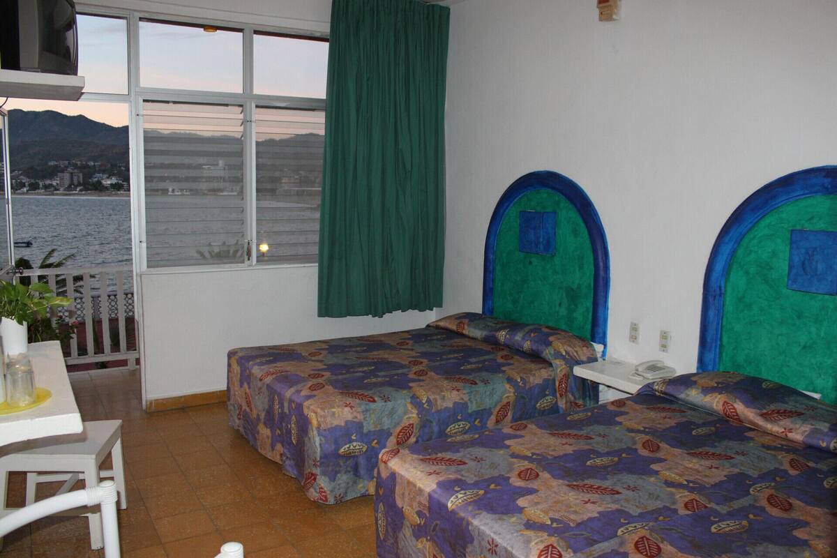 Ein Zimmer mit zwei Betten, einem Tisch, einem Stuhl, einem Fenster mit Meerblick und einem Vorhang.