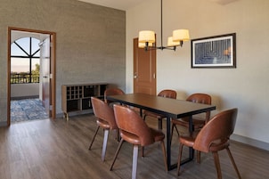 Un comedor con una mesa rectangular, seis sillas, una araña, una puerta y una obra de arte enmarcada en la pared.