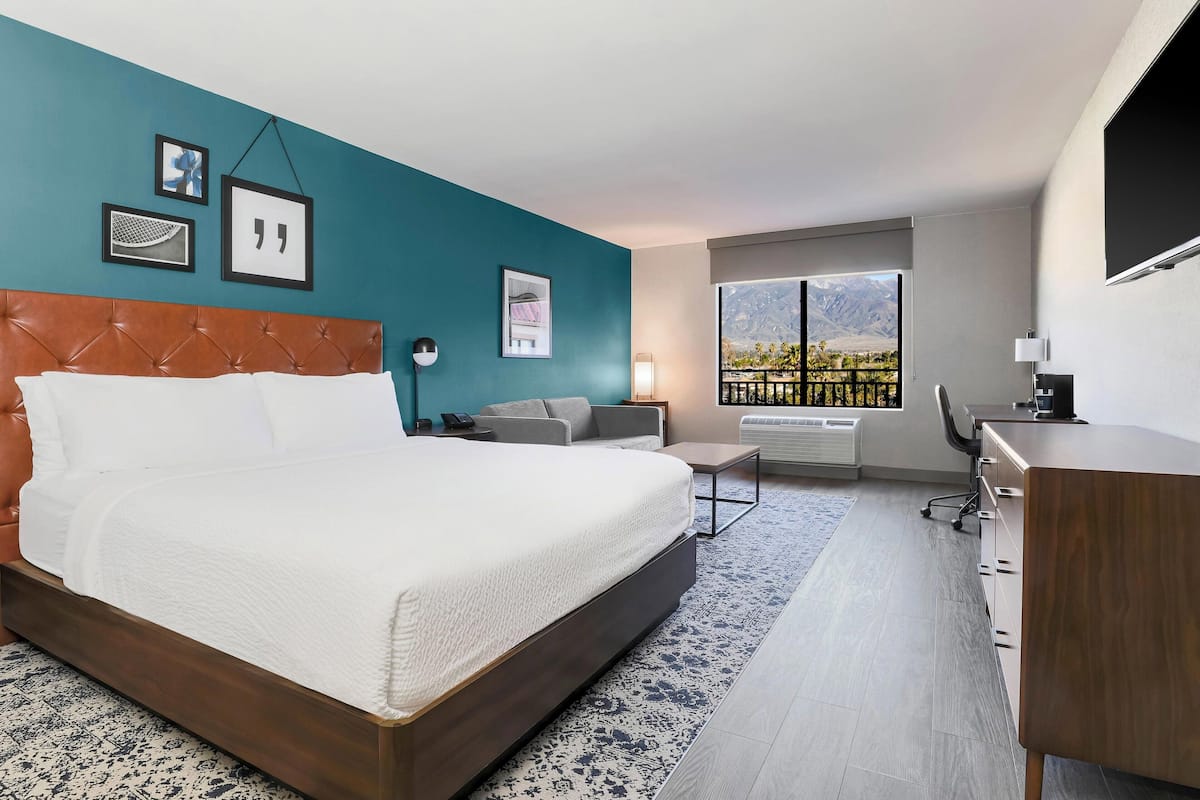 Ein Hotelzimmer mit einem großen Bett, einem Schreibtisch, einem Stuhl und Blick auf Berge durch das Fenster.