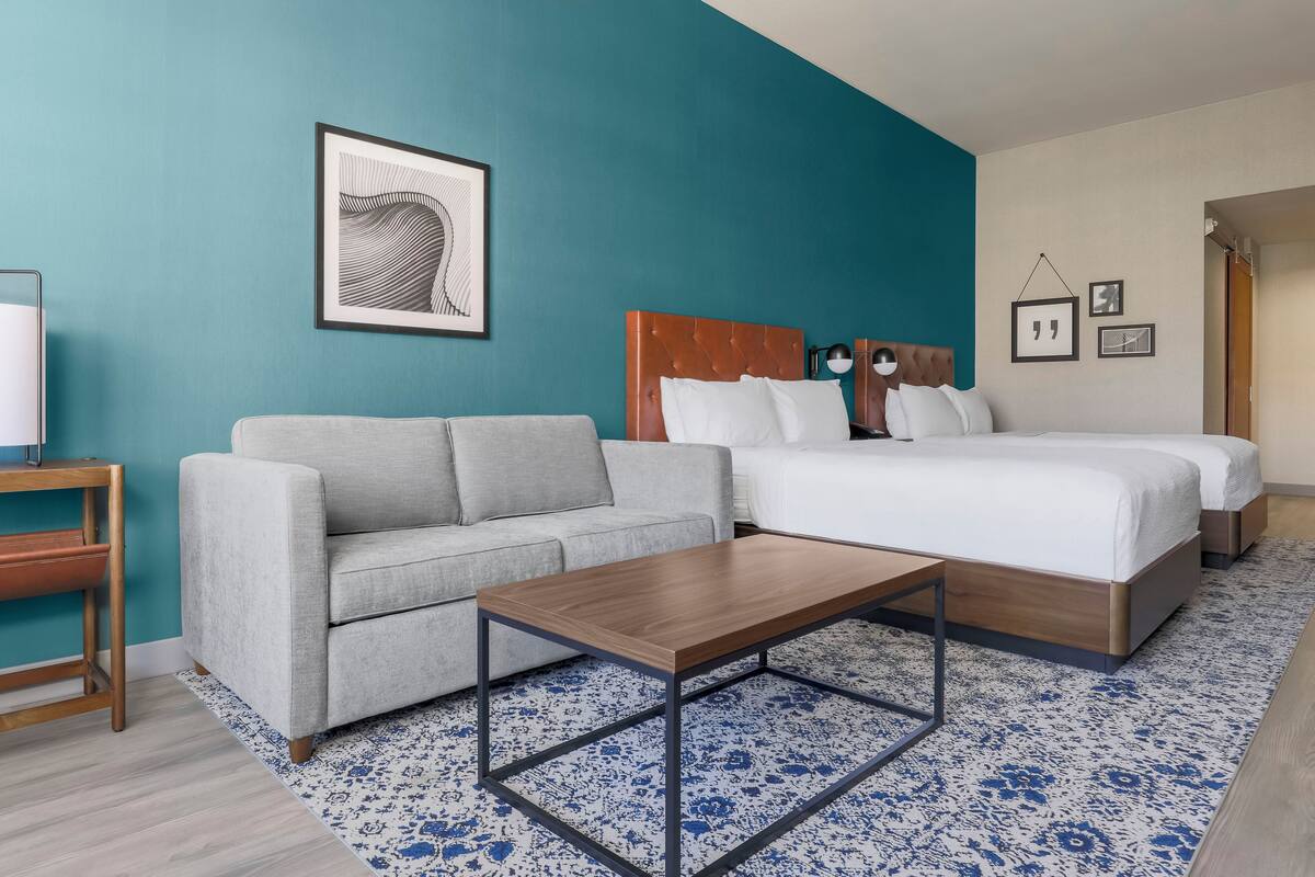Ein modernes Hotelzimmer mit einer Türkis-farbenen Akzentwand, einem grauen Sofa, einem hölzernen Bettgestell und einem gemusterten Teppich.