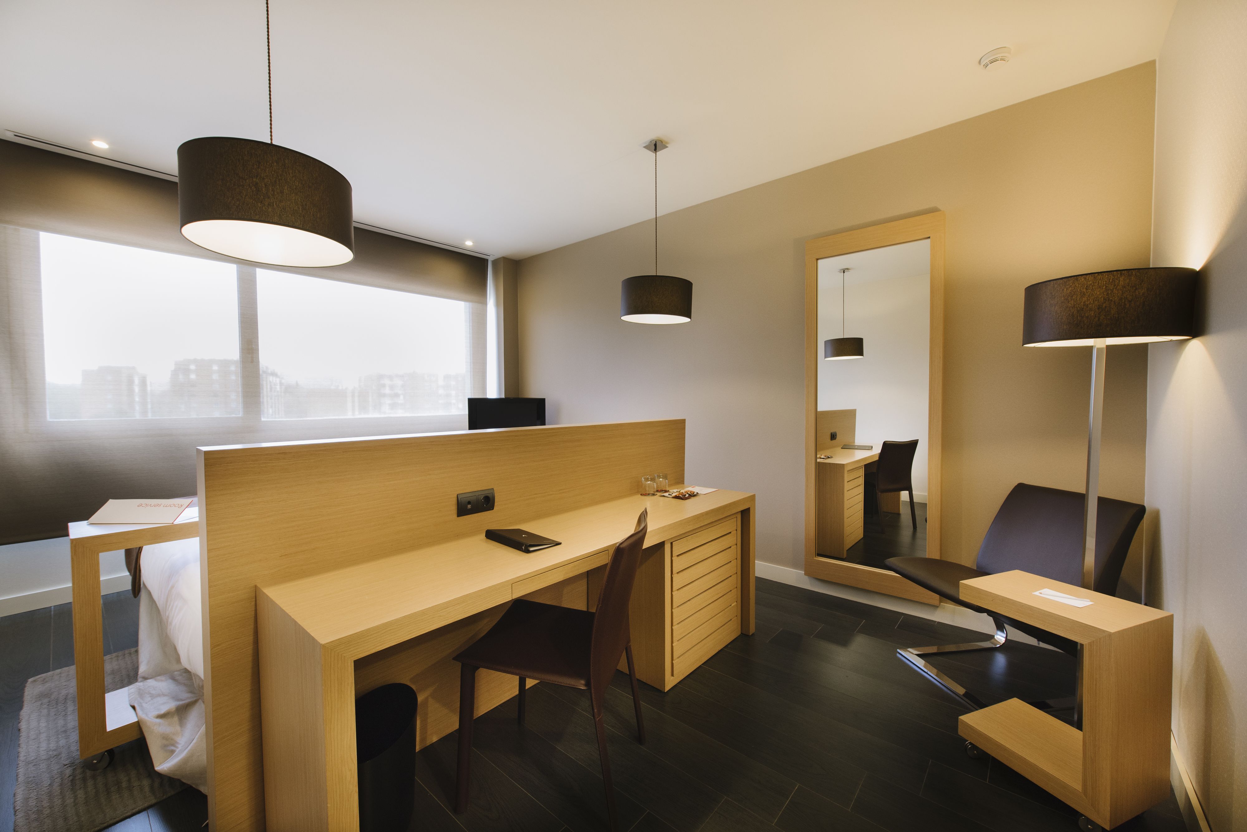 Une chambre d’hôtel moderne avec un bureau en bois, une chaise, un petit canapé, un grand miroir et deux lampes suspendues.