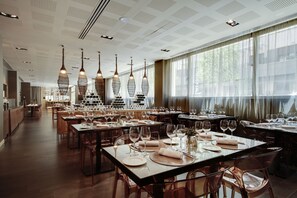 A restaurant with multiple dining tables set with white tablecloths and wine glasses, large windows with sheer curtains, and hanging pendant lights.