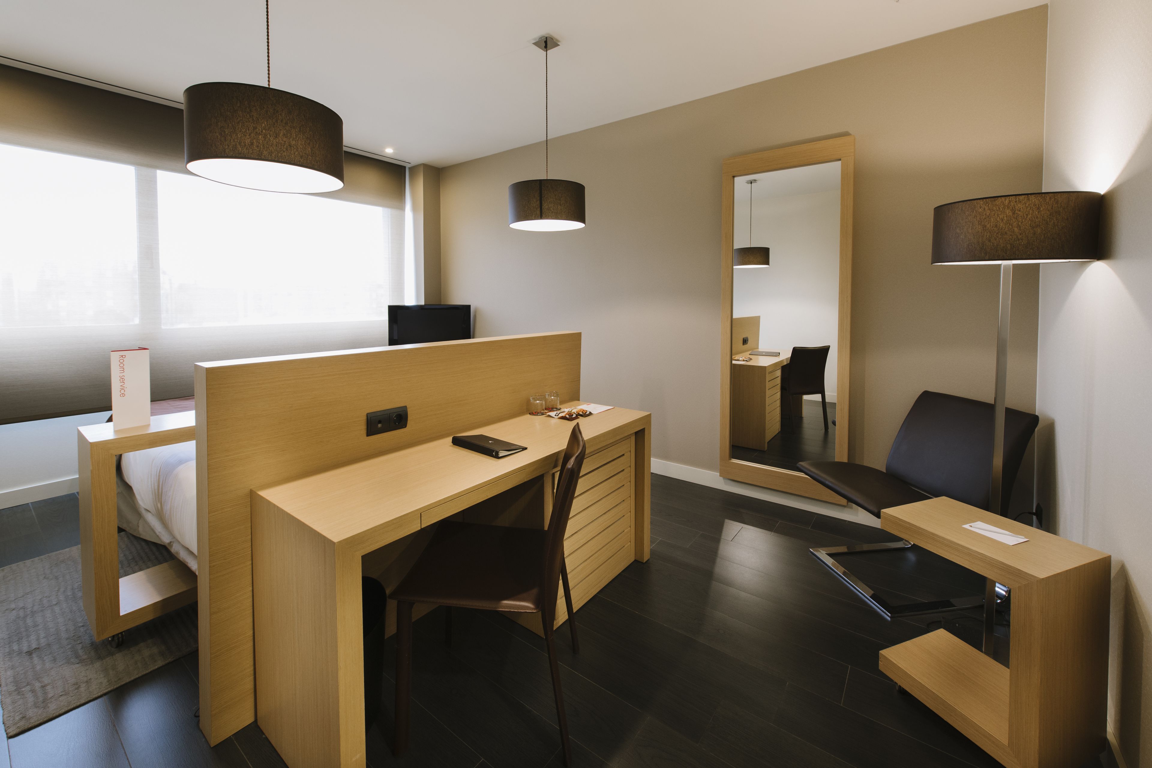 Una habitación de hotel moderna con un escritorio de madera, una silla, una cama, una lámpara de escritorio y un espejo grande.