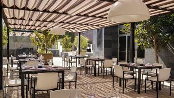 Un área de comedor al aire libre con mesas y sillas, cubierta por un techo a rayas y rodeada de follaje.