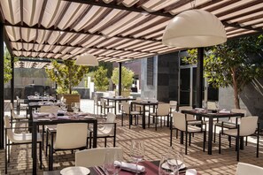 An outdoor dining area with tables and chairs, covered by a striped roof, and surrounded by greenery. - PCM Forum Alcalá (Alcala de Henares)