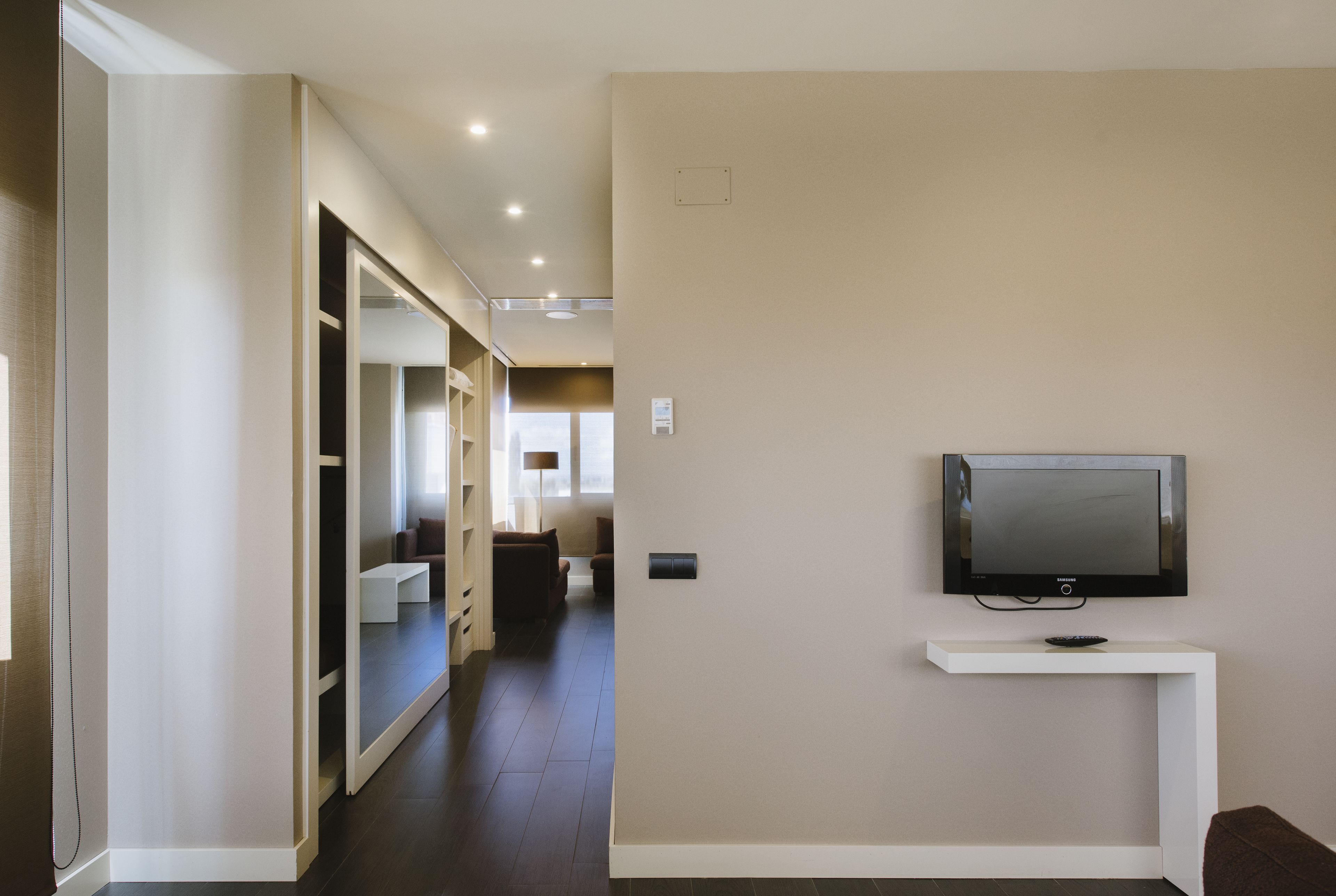 Un pasillo moderno de hotel con un televisor montado en una consola blanca, que conduce a una zona de estar con un sofá y una lámpara.