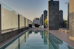 A modern outdoor pool area with clear glass walls, lounge chairs, and a view of surrounding buildings. - PCM Forum Alcalá (Alcala de Henares)