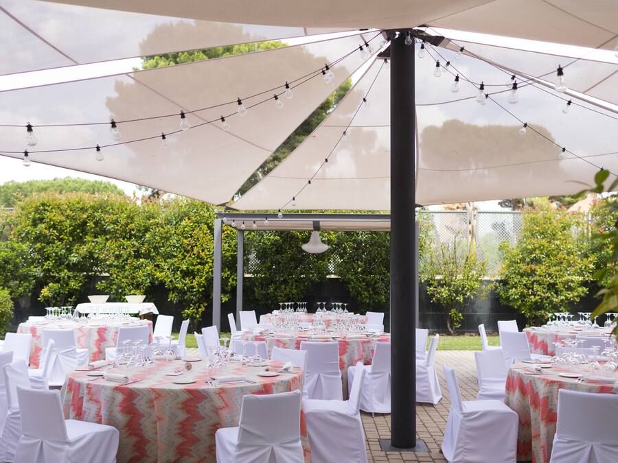 An outdoor event space with multiple round tables set up under large umbrellas, each table equipped with white tablecloths, chairs, and glassware.