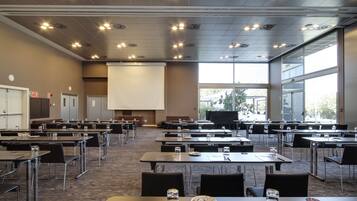 Un salón de conferencias con varias mesas y sillas, una pantalla de proyector y ventanas amplias.