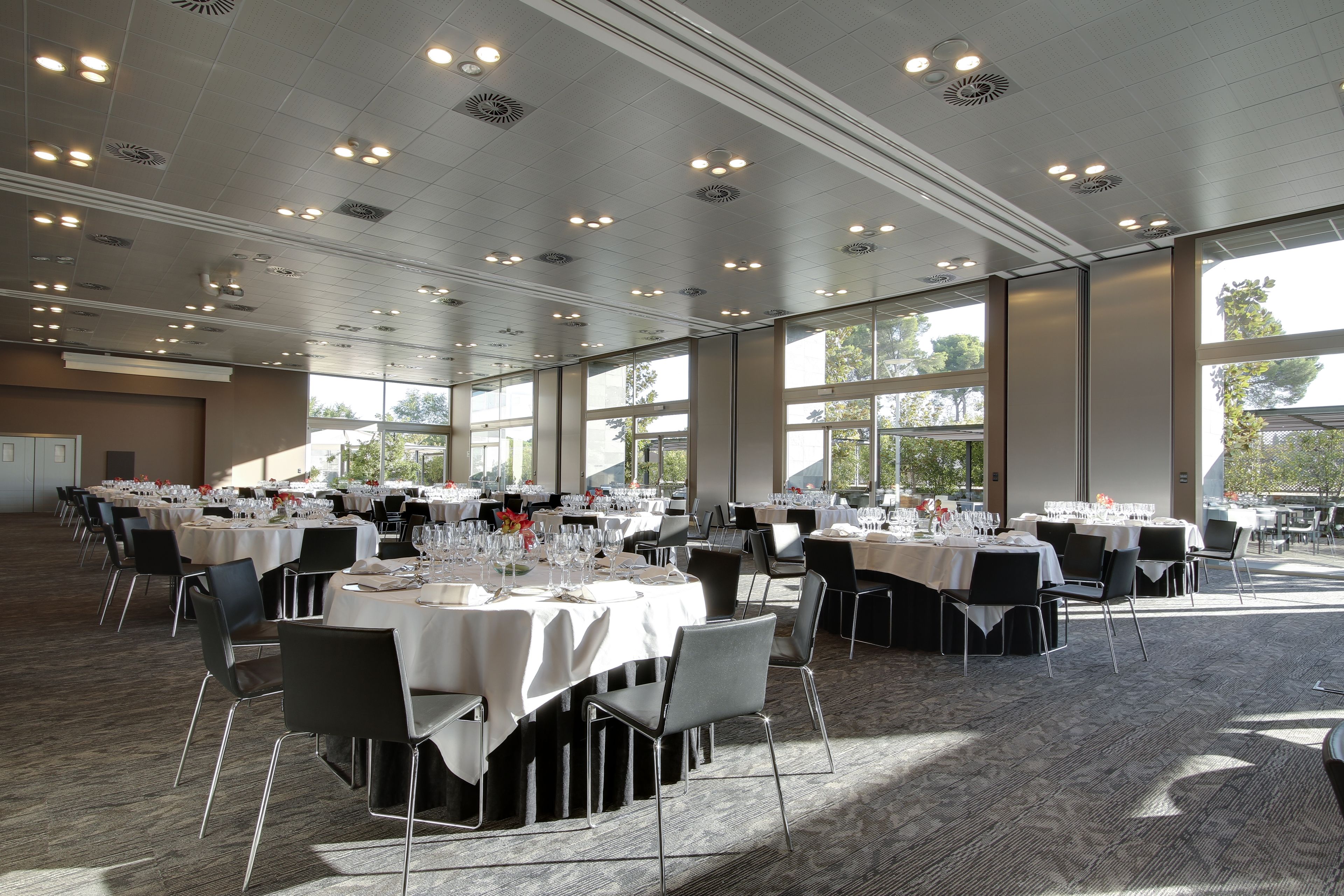 Una sala banchetti spaziosa con numerosi tavoli rotondi apparecchiati per la cena, ampie finestre che lasciano entrare luce naturale e un soffitto alto con illuminazione a incasso.