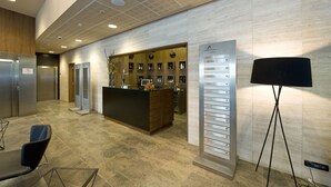 Un espace d’accueil d’hôtel comprenant un comptoir de réception en bois, un panneau d’affichage numérique et un coin salon avec des chaises.