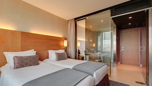 Une chambre d’hôtel avec deux lits, une cloison en verre, une tête de lit en bois et une porte coulissante donnant sur une salle de bain.