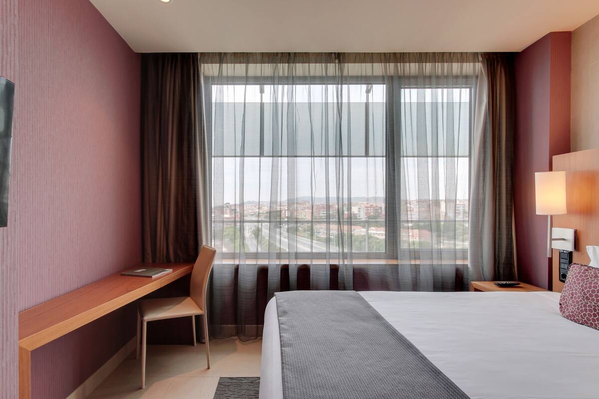 Ein Hotelzimmer mit Bett, Schreibtisch, Stuhl, Nachttisch und einem großen Fenster mit Vorhängen.