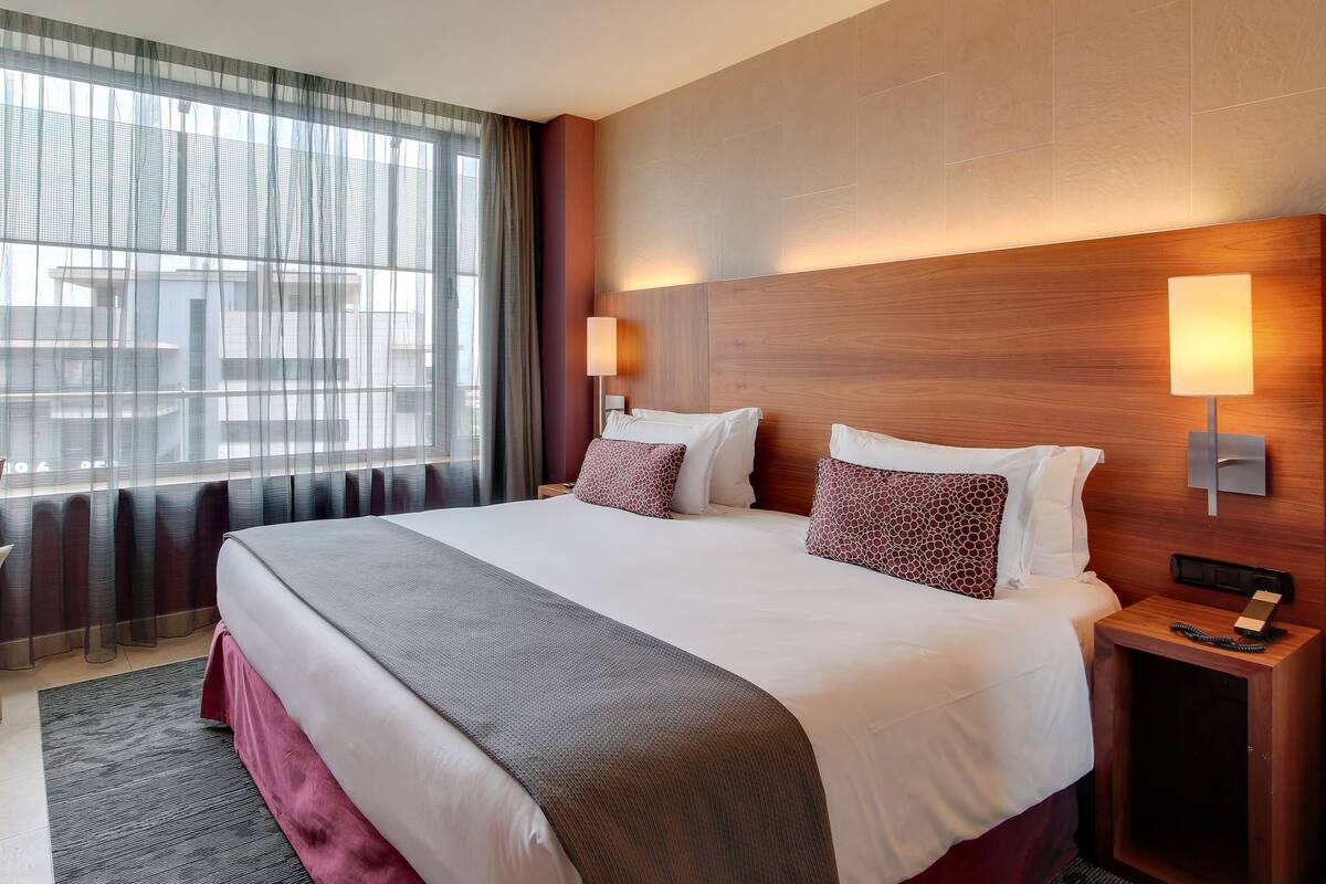 Ein Hotelzimmer mit einem großen Bett, zwei Nachttischlampen, einem Holz-Kopfende, einem Schreibtisch mit Stuhl und Blick durch durchscheinende Vorhänge.