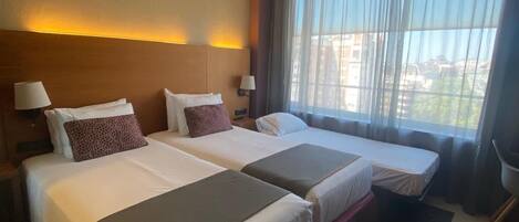 A hotel room with two beds, a large window with sheer curtains, and a view of buildings outside.