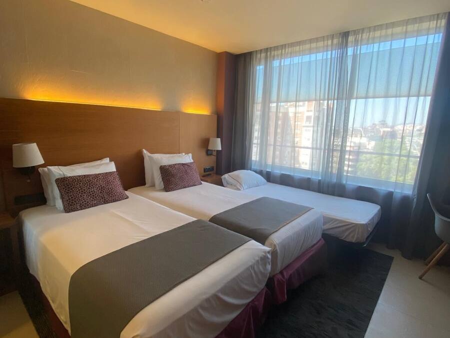 A hotel room with two beds, a large window with sheer curtains, and a view of buildings outside.