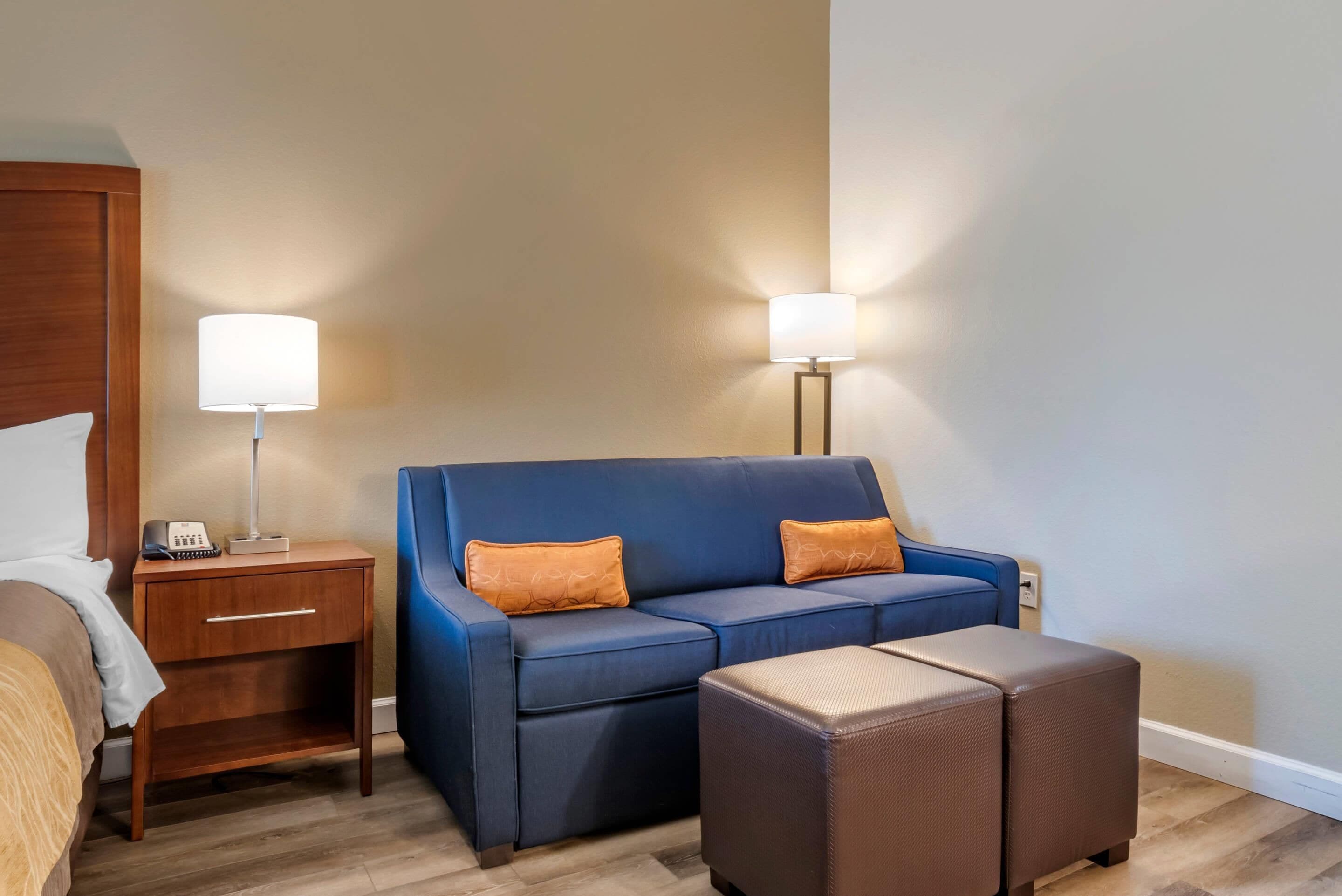 A hotel room with a bed, a blue sofa with two orange cushions, a small wooden nightstand with a lamp, and a brown ottoman.