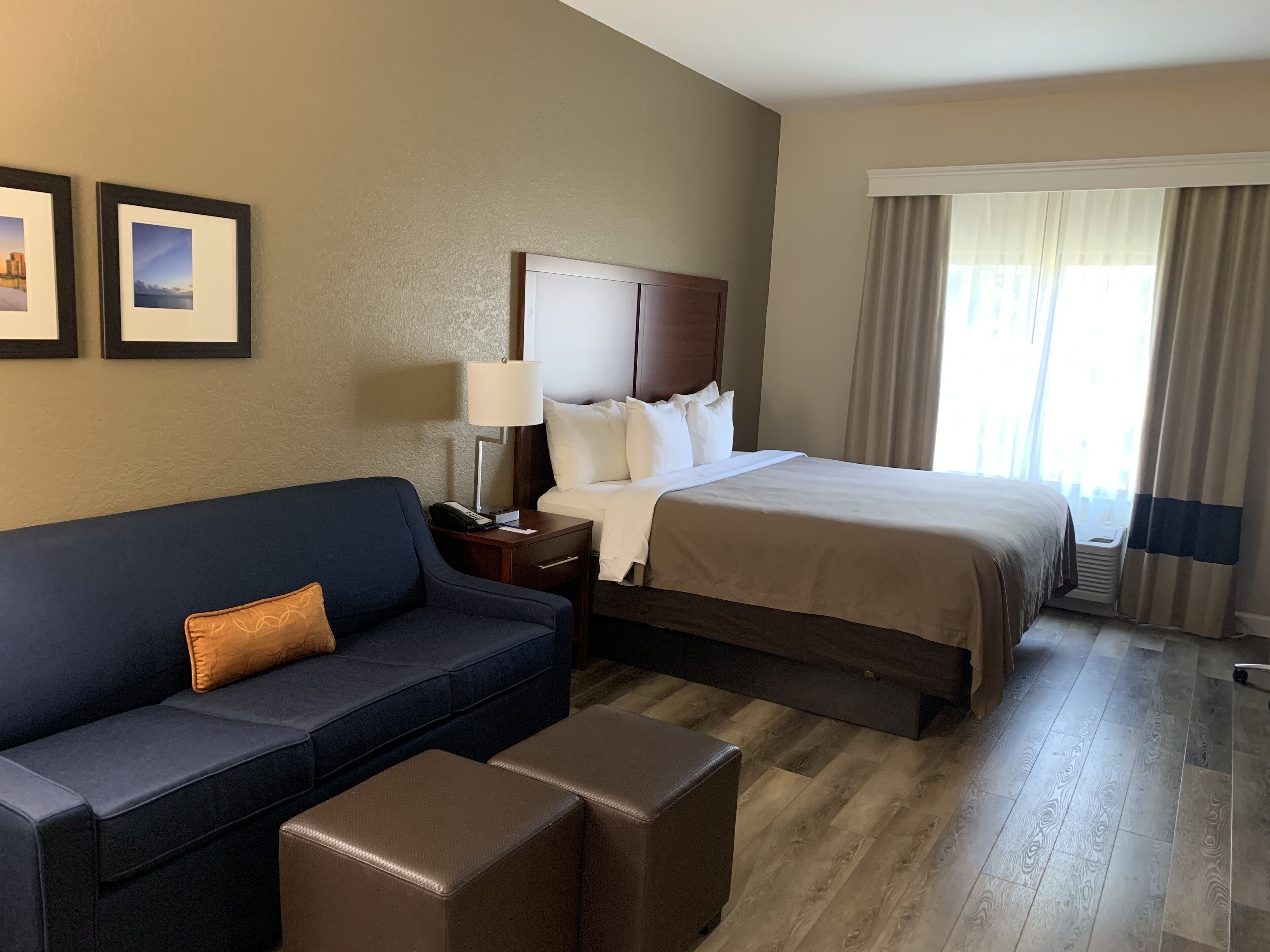Quarto de hotel com cama, sofá, mesa de cabeceira, luminária e janela com cortinas.