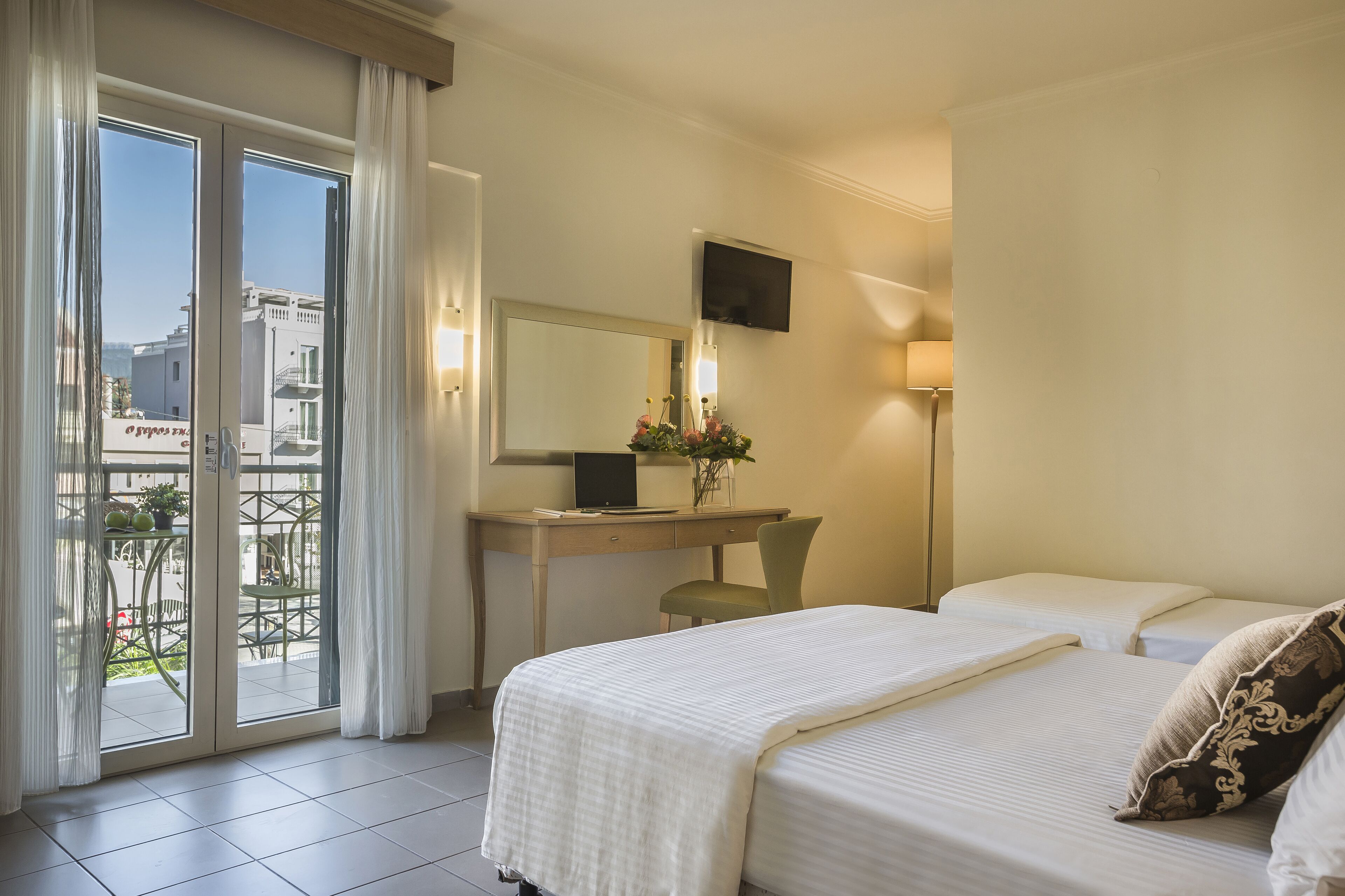 A hotel room with a bed, a desk with a computer, a chair, a TV, and a balcony with curtains.