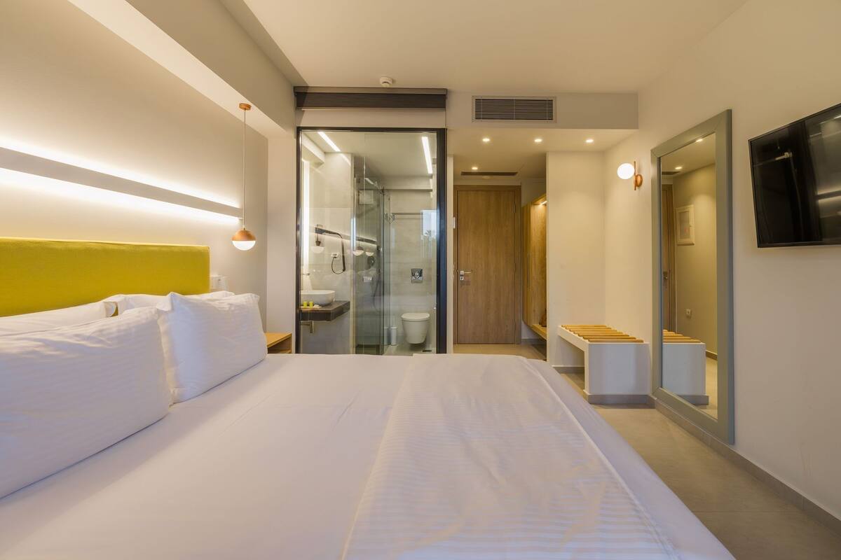 Una habitació d'hotel moderna amb un llit gran, un bany amb plat de dutxa de vidre i un armari de fusta.
