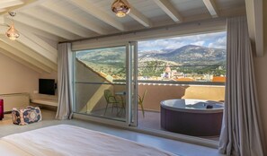 A bedroom with a large bed, a sliding glass door leading to a balcony, a hot tub, and a view of mountains and buildings. - Ionian Plaza Hotel & Spa (Kefalonia)