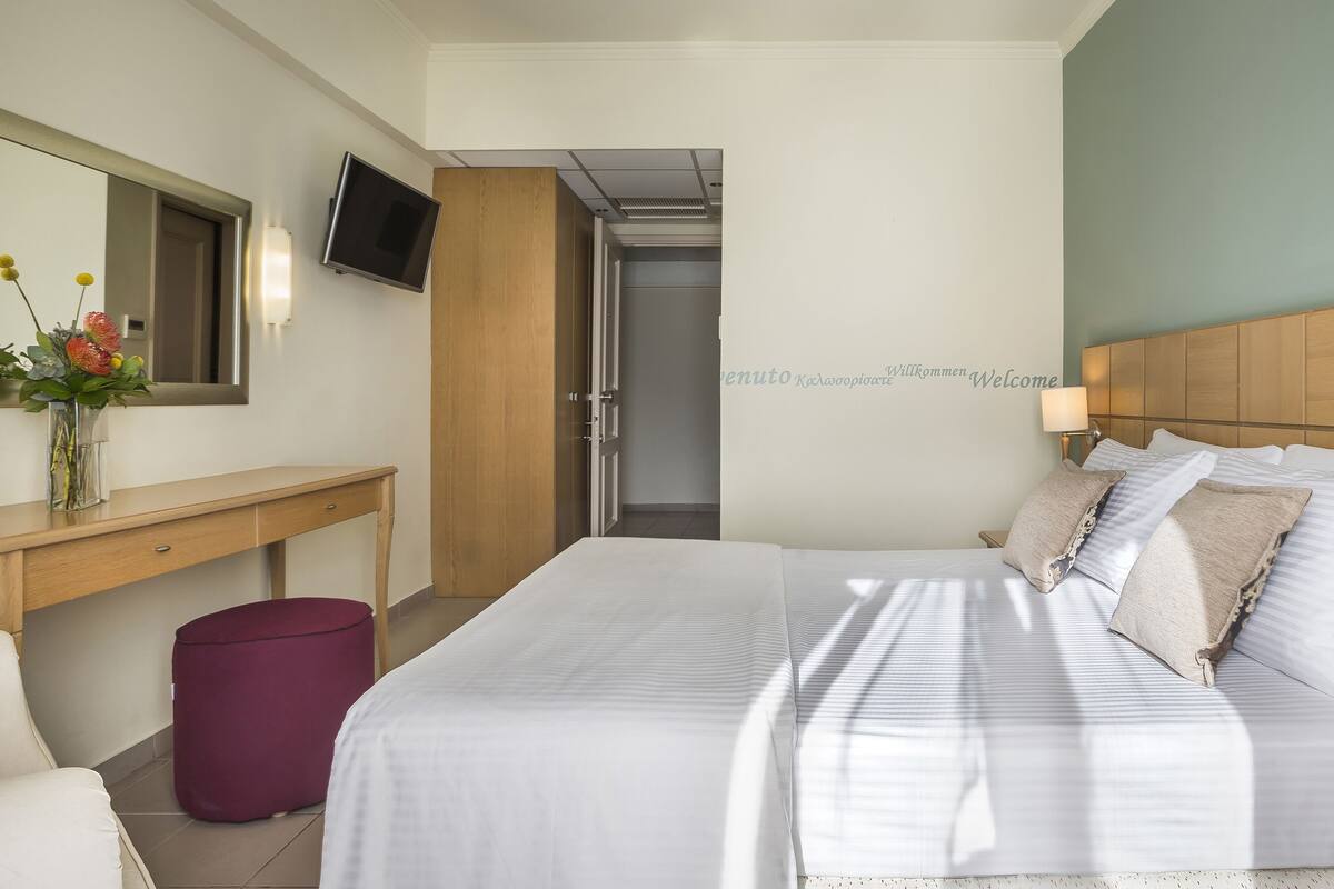 Habitació d'hotel amb un llit, un escriptori, un mirall, una televisió i una paret amb un missatge de benvinguda.