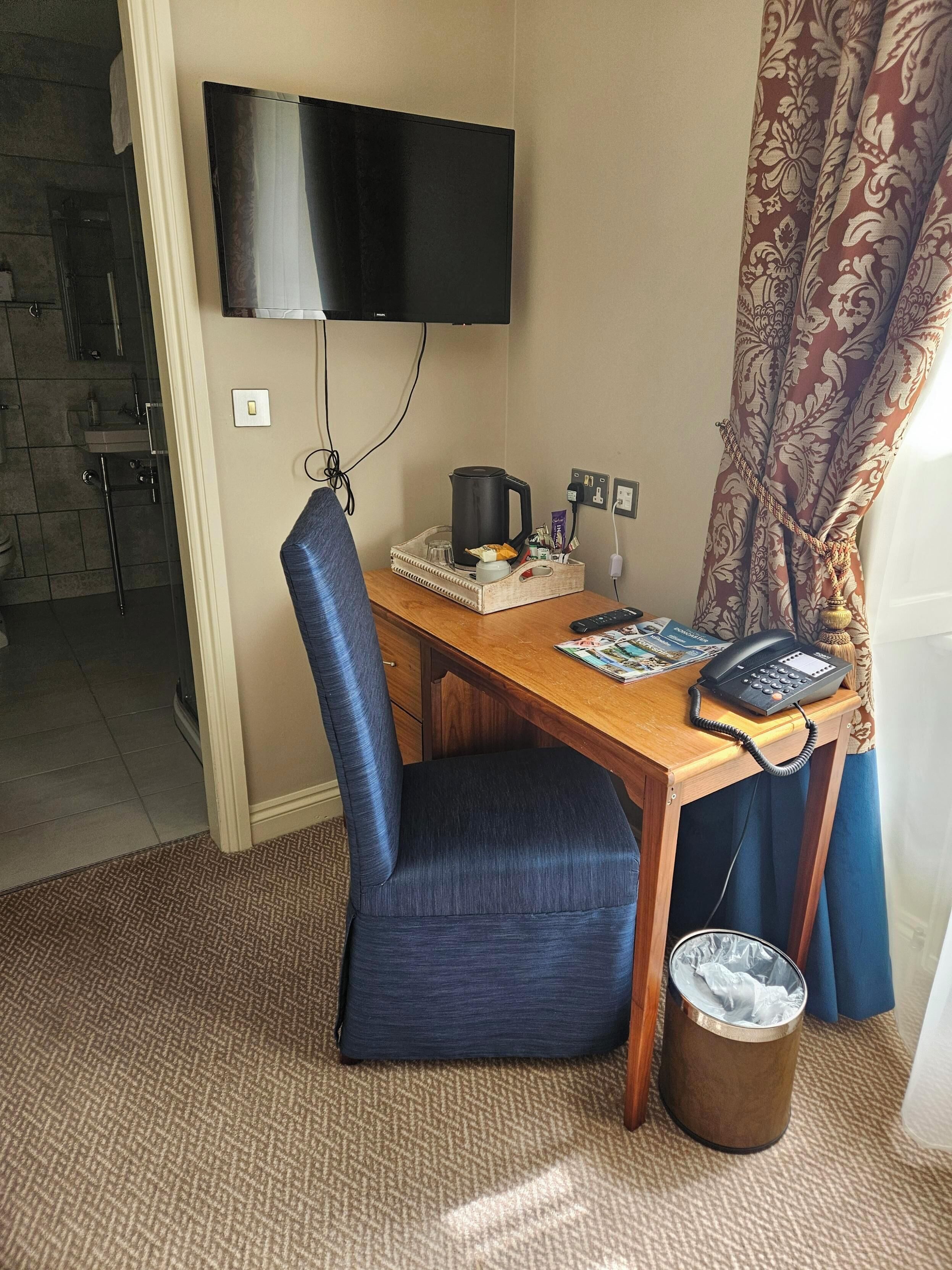 Quarto de hotel com uma escrivaninha de madeira, uma cadeira azul, uma televisão na parede, uma cafeteira, um telefone e uma lixeira.