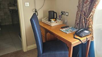 Quarto de hotel com uma escrivaninha de madeira, uma cadeira azul, uma televisão na parede, uma cafeteira, um telefone e uma lixeira.