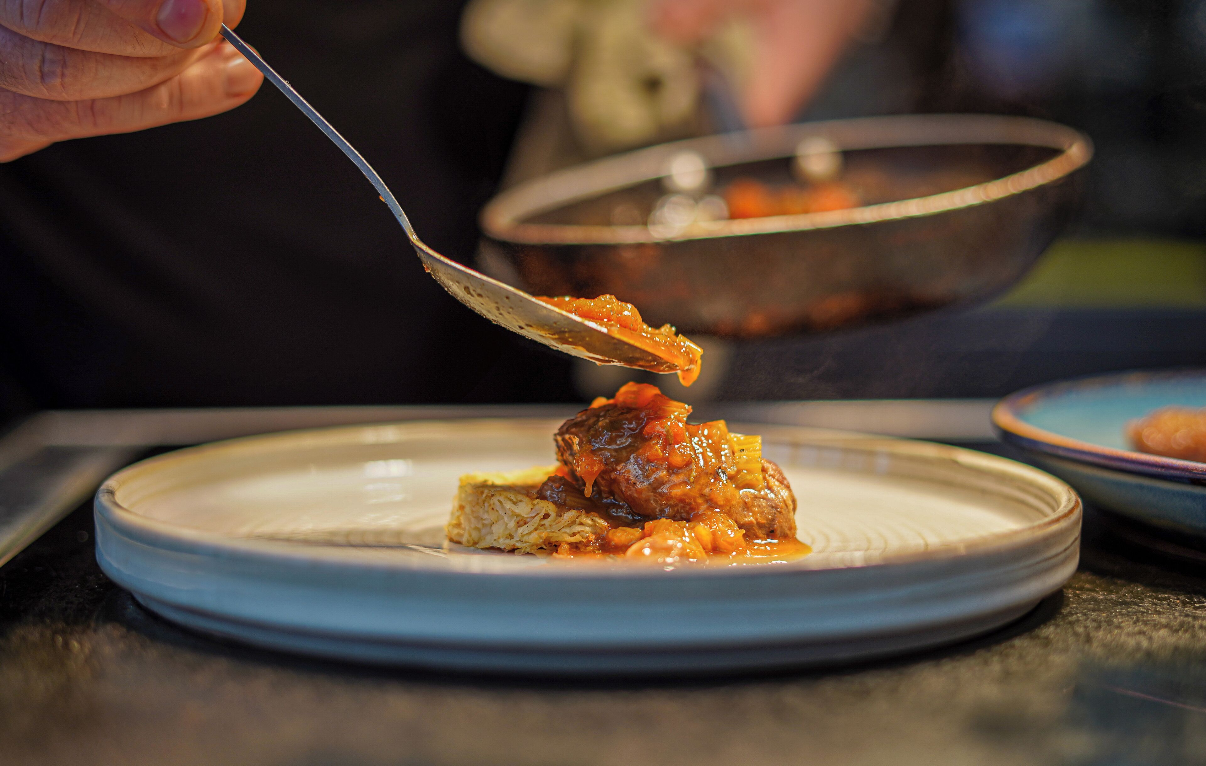 A spoon lifting food from a plate, with a blurred bowl in the background.