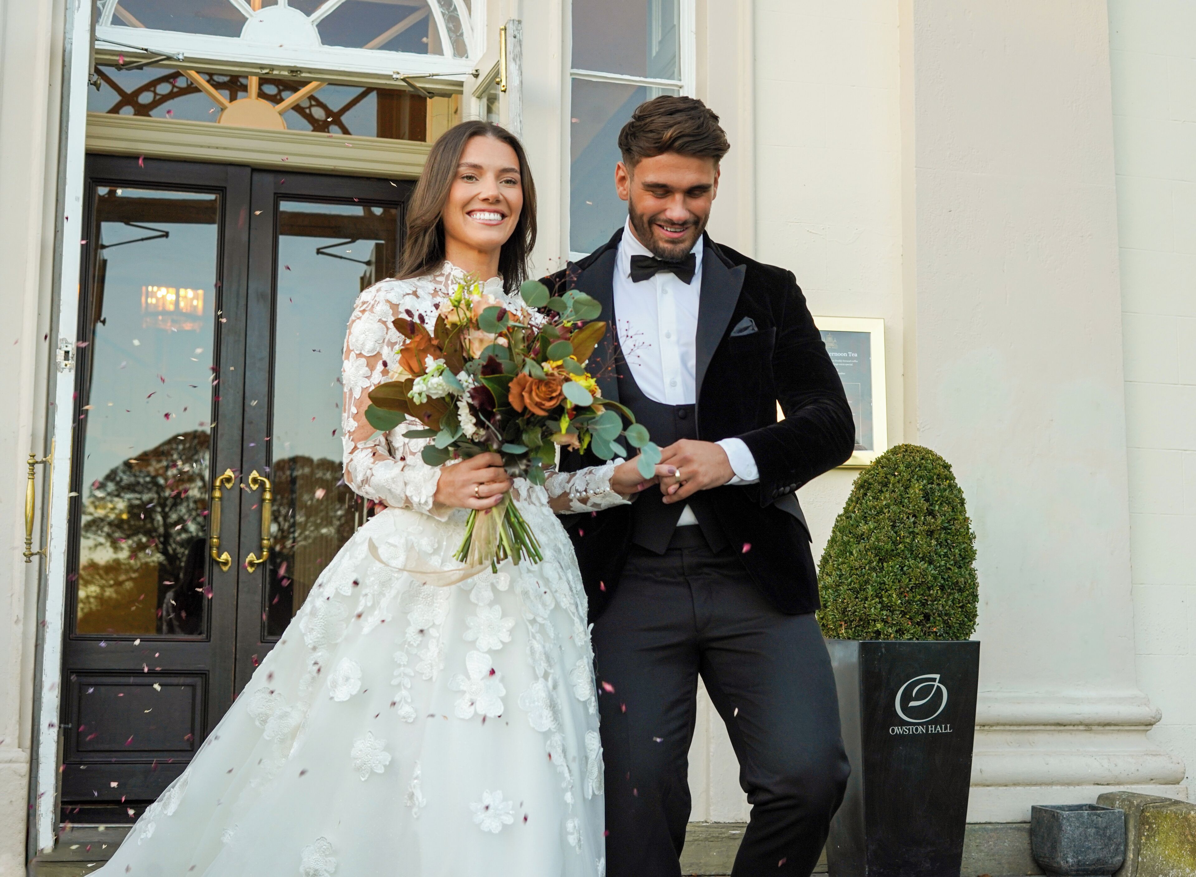 Ein Brautpaar in Hochzeitskleidung steht vor einem Gebäude mit einer schwarzen Tür und einem Schild, auf dem 'OWSTON HALL' steht.