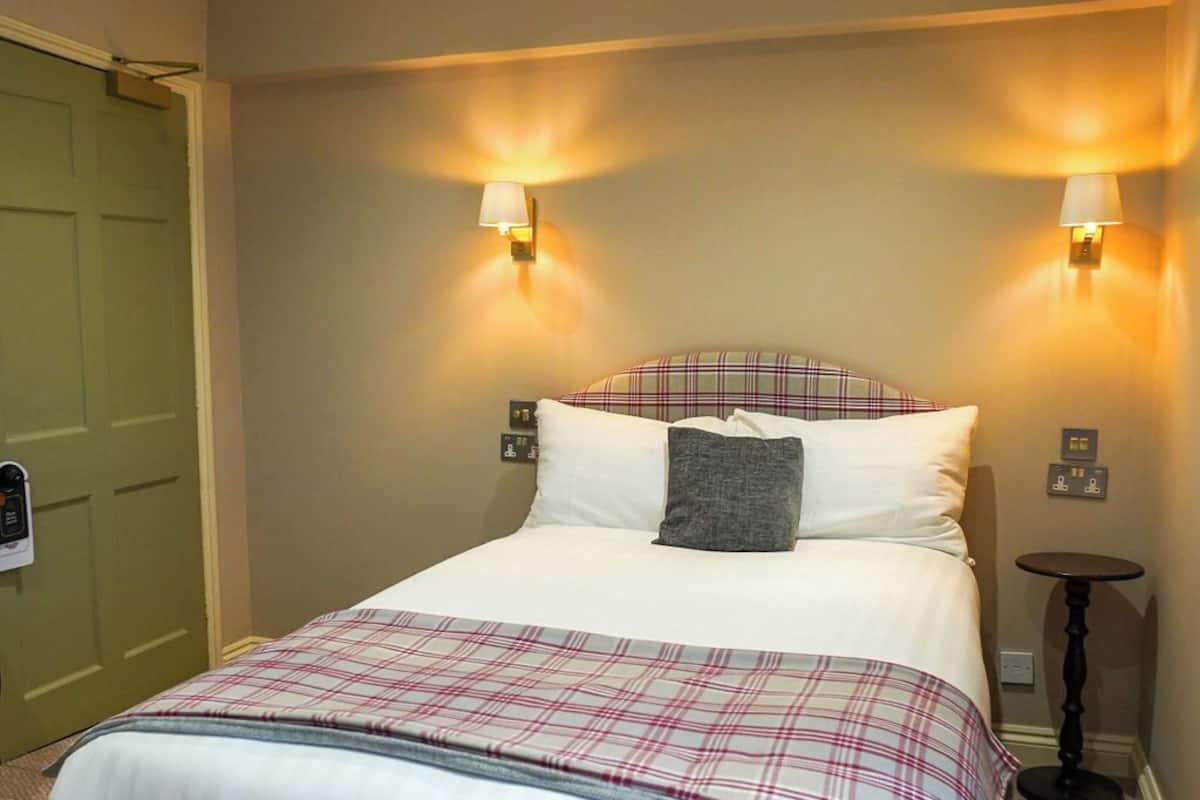 A hotel room with a large bed, two wall-mounted lamps, a small round table, and a green door.