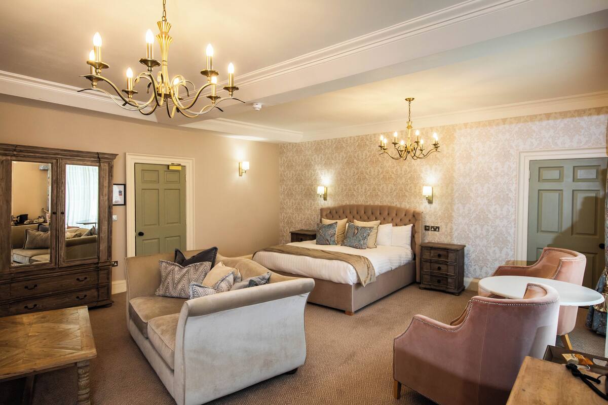 A spacious bedroom with a large bed, a sitting area with a sofa and armchairs, a wooden dresser, and a chandelier.
